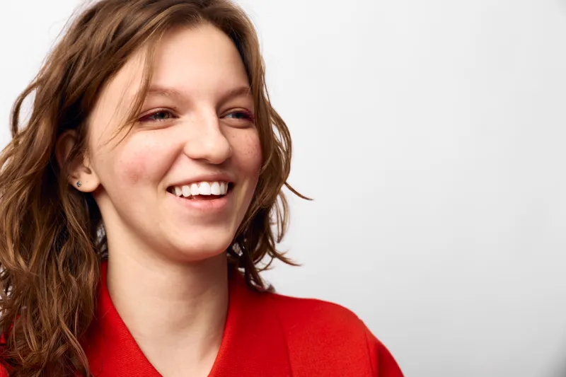 Smiling woman with wavy brown hair wearing a red sweater looks away from camera on white background. Concept of happiness, casual style, natural beauty, and positive emotions.