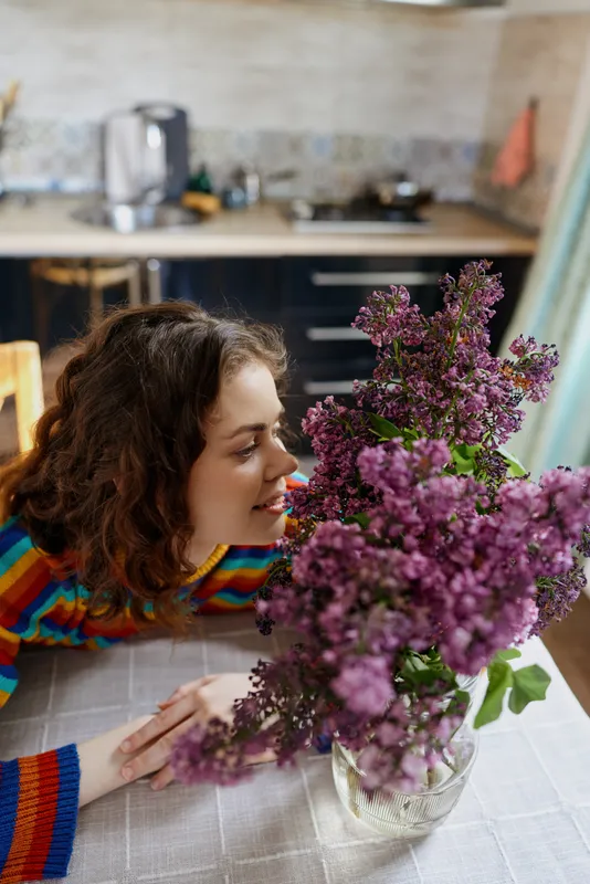 A young woman with curly hair leans over a purple lilac bouquet on a bright kitchen table, inhaling its scent with a gentle, contemplative smile and warm morning light.