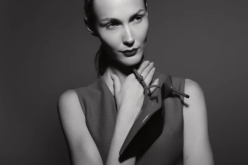 Portrait of a pale white woman holding a high heel shoe near her neck, wearing sleeveless top in monochrome. Concept for fashion, elegance, and style with dramatic lighting.