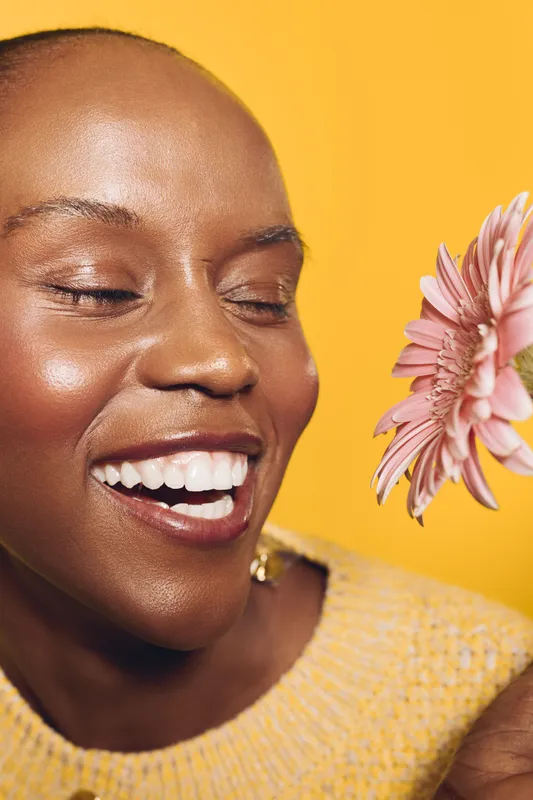 Smiling dark skinned African woman with shaved head holding pink flower near face on yellow background. Concept of natural beauty, happiness, skincare, and joyful female portrait.