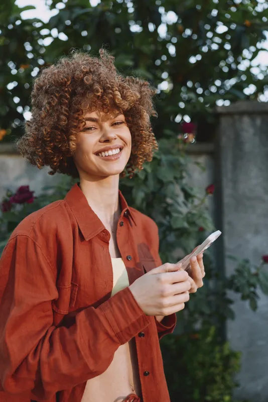 Young woman with curly hair smiles while holding a knife in an outdoor setting, a candid moment of everyday confidence and bold style