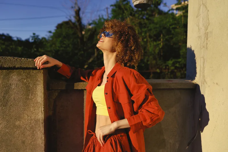 Woman stands outdoors, leaning on a wall, wearing a red jacket and yellow top, sunglasses, basking in sunlight. Lifestyle concept focusing on fashion, confidence, and relaxed everyday moments