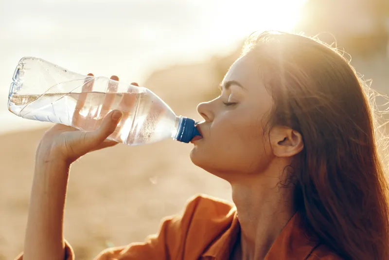 Woman drinking water outdoors in sunlight, calm face, healthy wellness lifestyle, hydration moment, close up profile, clear bottle, natural background, relaxed posture, fresh breeze