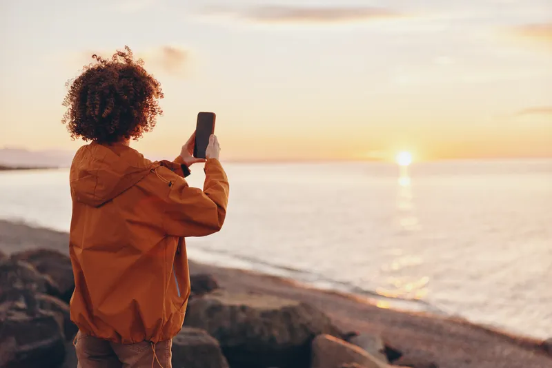 Woman lifestyle, seaside sunset scene, curly haired woman on rocky shore, holding a phone for a selfie, warm golden light, ocean waves in background, peaceful travel mood