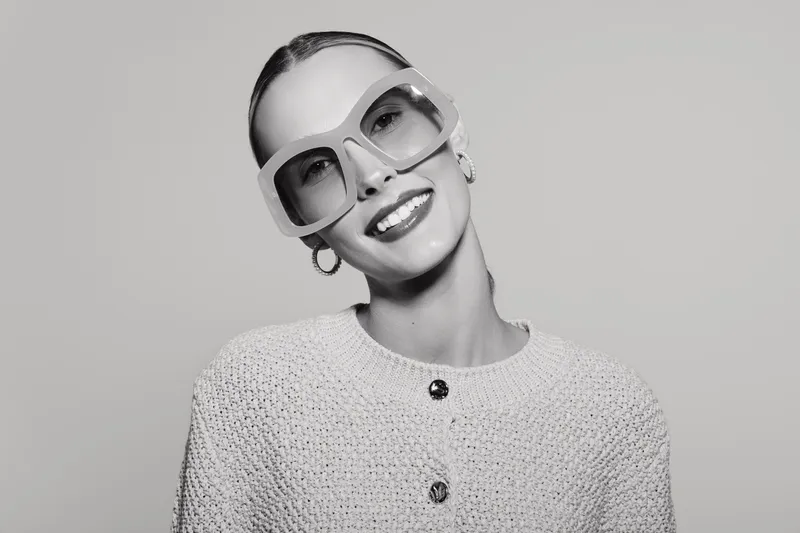 Smiling white woman with pale skin and slicked back hair wears large sunglasses and knitted sweater. Studio portrait in black and white with fashion and beauty concept.