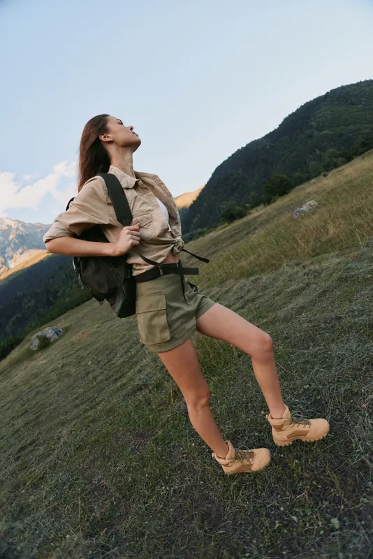 Woman with a backpack stands on a hillside, gazing up at the sky. Casual hiking attire, outdoor adventure, nature exploration, and wellness lifestyle in a scenic landscape.