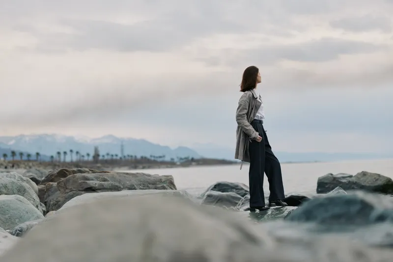 Woman in casual clothes stands on rocky shore looking at distant mountains and cloudy sky. Concept of contemplation, nature, and outdoor lifestyle with calm mood.
