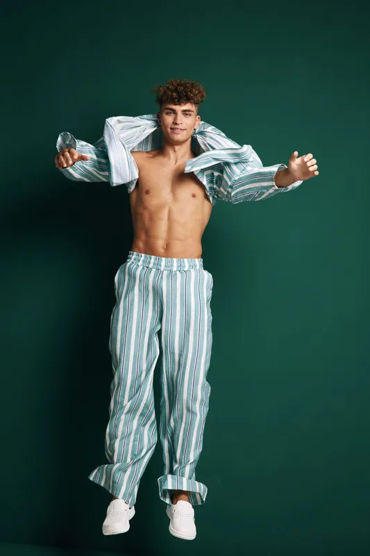 Young man jumping with striped pajamas open shirt and bare chest on dark background. Casual style, energetic pose, smiling male model with curly hair and white sneakers in studio.