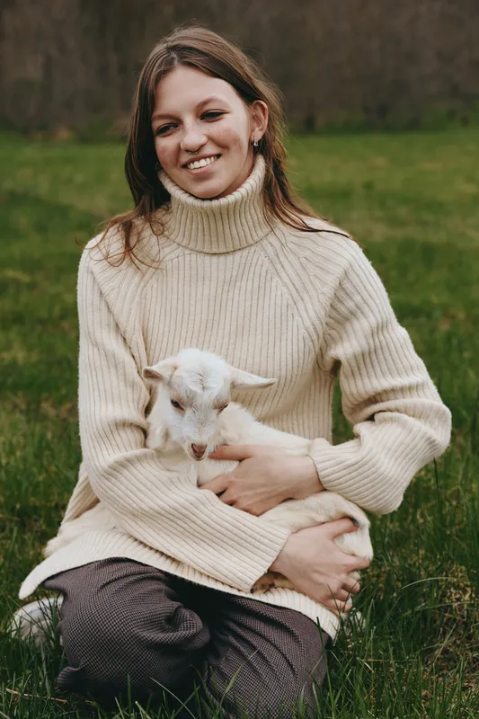 Smiling woman in a cream turtleneck sits on grass cradling a young goat kid, sunlit meadow scene conveying gentle lifestyle, animal companionship, and rustic wellness