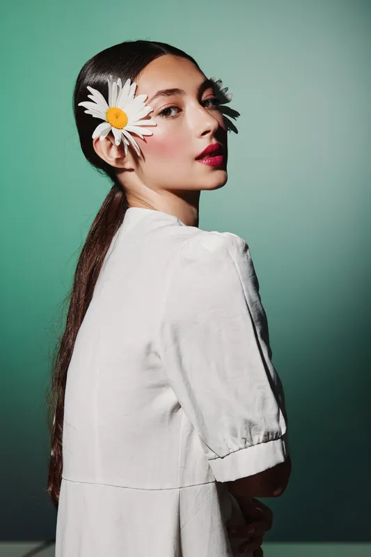 Creative fashion portrait of young woman with long hair and daisies in her ears. She wears a white outfit against a clean pastel green background. Beauty concept with floral accent.