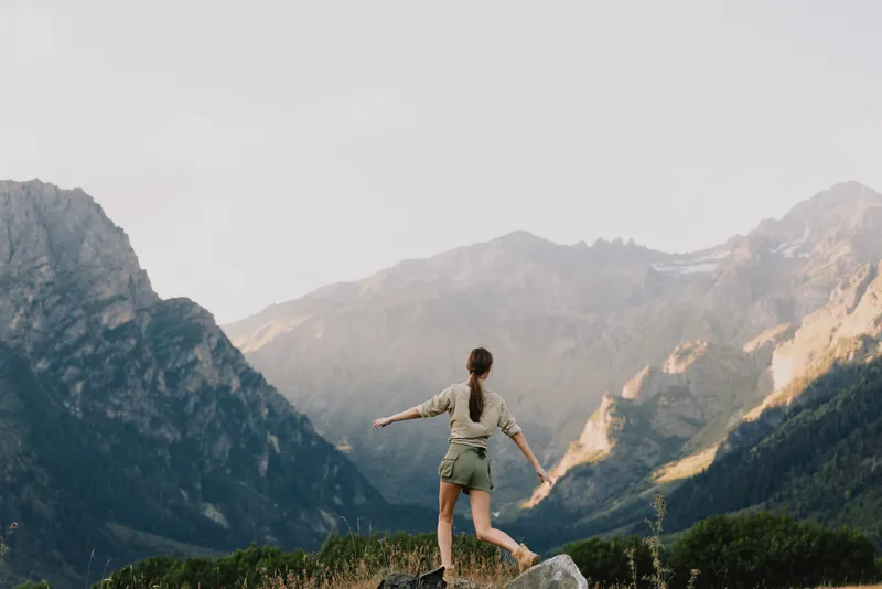 A woman balances on a rocky overlook with arms extended against a dramatic mountain panorama, embodying a moment of freedom and adventurous spirit in the outdoors.