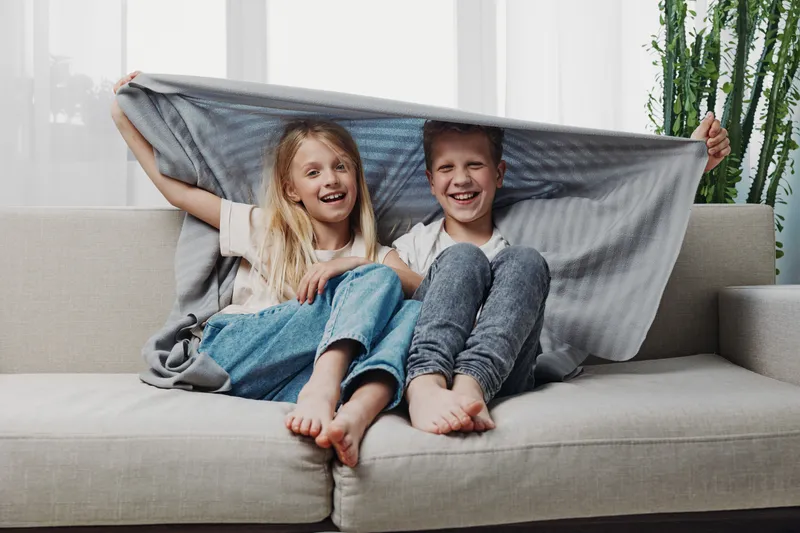 Happy children sitting on couch under grey blanket smiling and playing at home. Cozy family moment with barefoot boy and girl enjoying warmth and togetherness in bright living room.