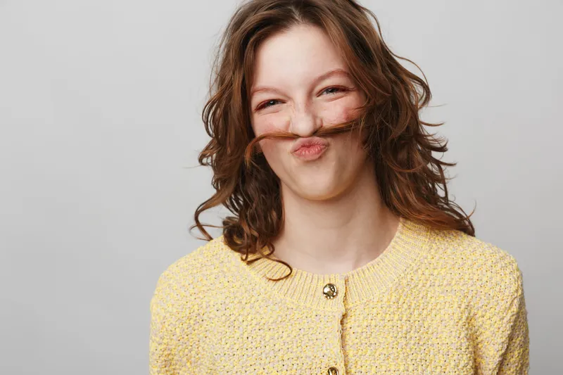 Young woman with curly brown hair makes funny face holding hair over upper lip like mustache. She wears yellow knitted sweater on light grey background. Concept of humor and playfulness.
