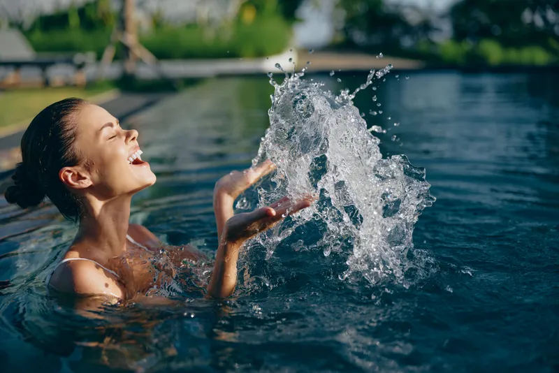 A smiling woman enjoys a playful splash in a sunlit outdoor pool, droplets arc in the air. A carefree lifestyle moment capturing joy, refreshment, and summer leisure.