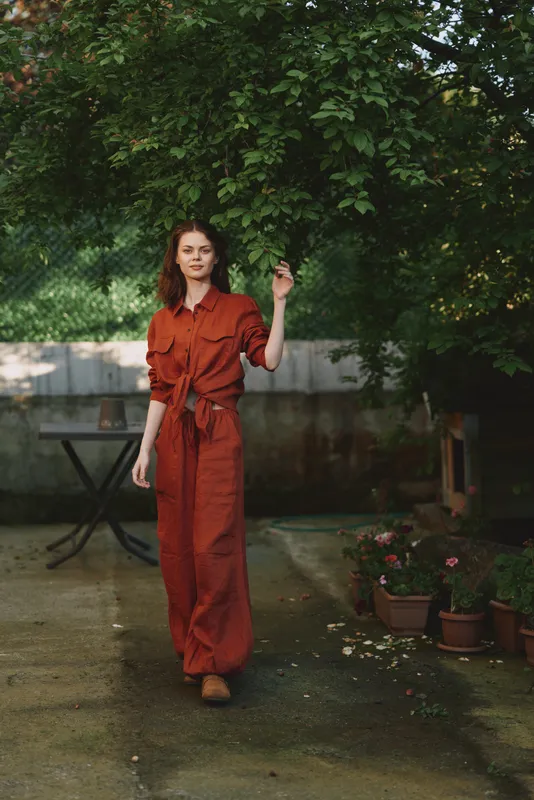 Cheerful woman lifestyle outdoors in a rust jumpsuit, posing in a leafy garden with a soft breeze, sunlit tones, casual elegance, relaxed posture, natural colors