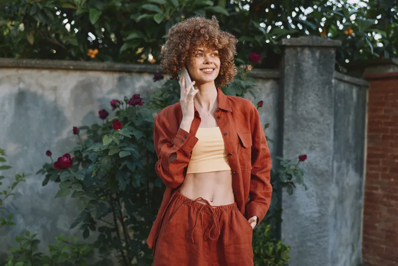 Smiling woman with curly hair wearing a rust orange jacket and yellow crop top. Outdoor garden backdrop with greenery and brick wall. Casual fashion, vibrant lifestyle, confident mood