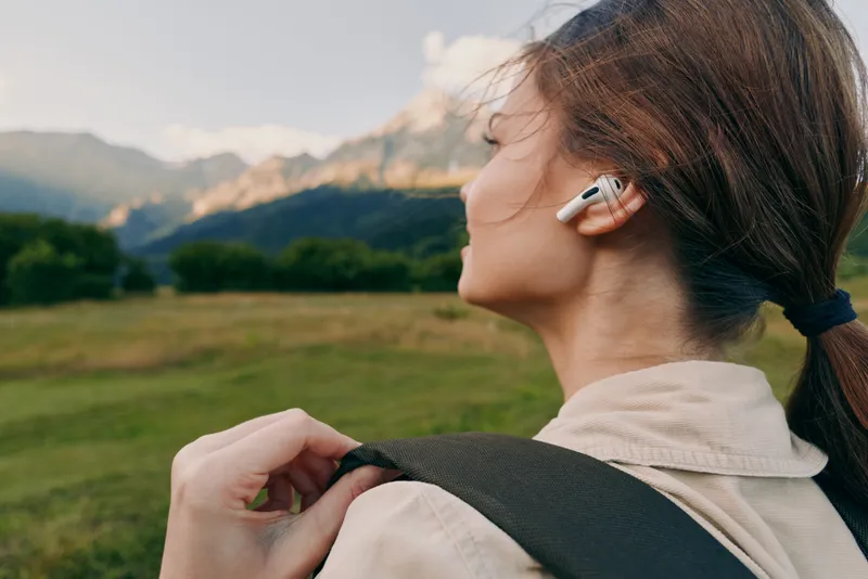 Woman outdoors with backpack looking over a green valley, wearing a light jacket, earbud in ear, sunny day, natural colors, relaxed lifestyle moment and quiet scenery in the background