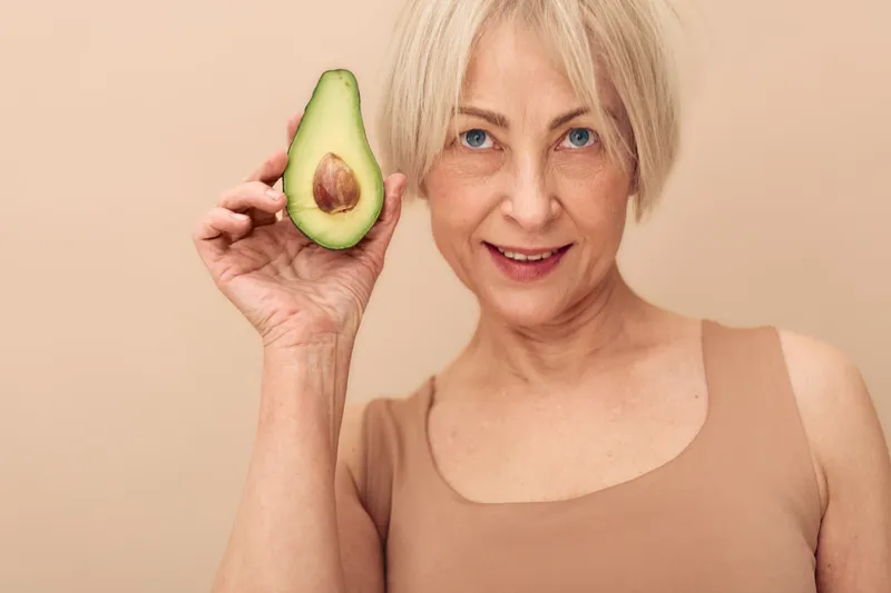 Smiling mature woman with short blonde hair holds half avocado near face on beige background. Concept of healthy lifestyle, nutrition, natural food, and wellness for older adults.