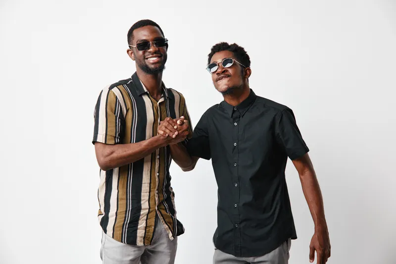 Two African American men with dark skin wearing sunglasses shaking hands and smiling in casual summer clothes on white background for friendship and connection.