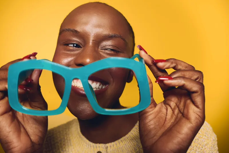 Happy smiling black woman with dark skin holds oversized blue glasses frame in front of her face on yellow background. Concept of joy, fun, and playful mood with vibrant colors.