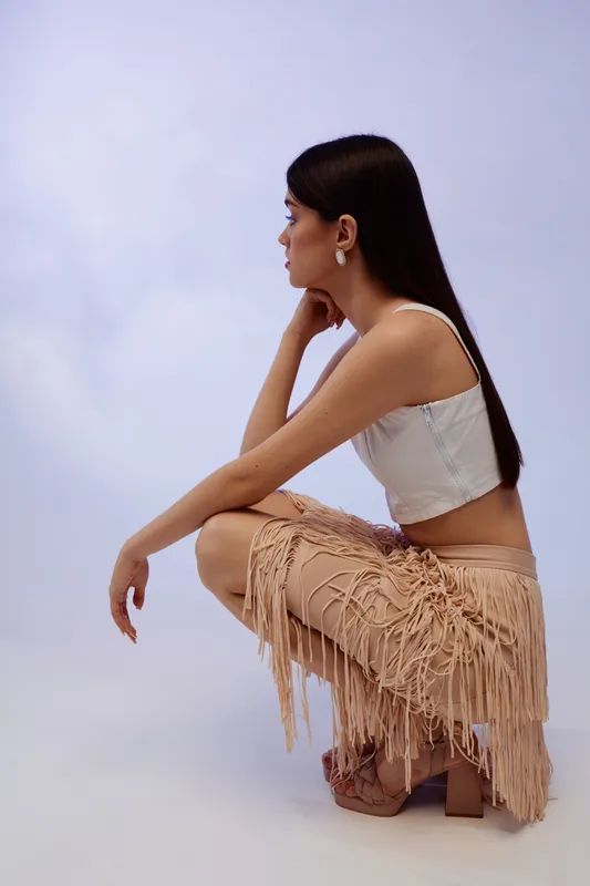 Young woman with long dark hair wearing a white top and fringed skirt crouches on a light background. Concept of woman beauty fashion and style in studio lighting.