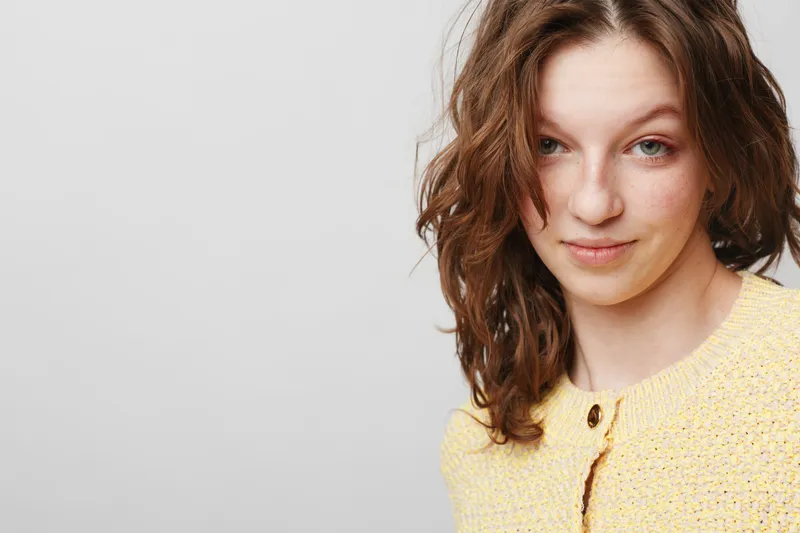 Young woman with wavy brown hair wearing a yellow knitted sweater looks confidently at the camera. Studio portrait with soft lighting on a plain light gray background.