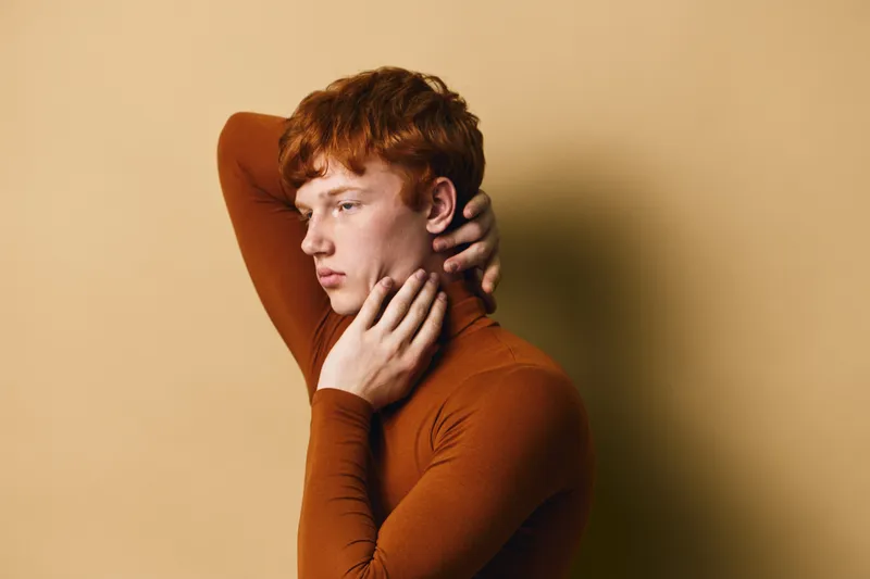 Portrait of red hair man wearing brown turtleneck sweater posing with hand on neck against beige background. Young male model with thoughtful expression and pale skin.