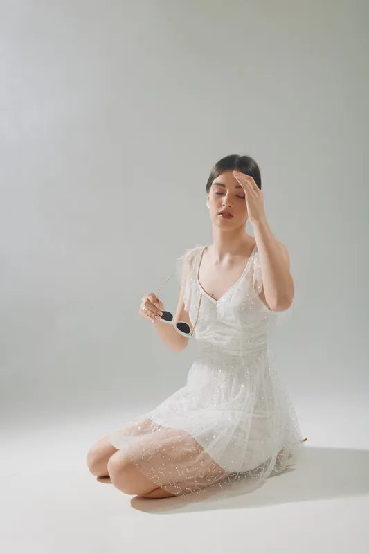Young woman in a white dress holding sunglasses with closed eyes. Soft natural light highlights her beauty and fashion style in a minimalistic studio setting.