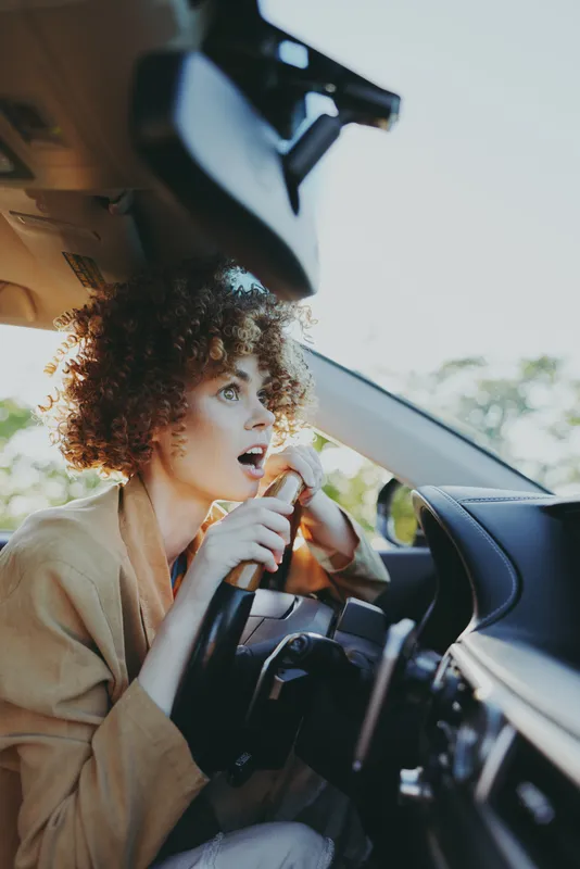 Cheerful woman driver, surprised expression, steering wheel closeup, sunlit car interior, warm retro tones, casual lifestyle moment, natural light, candid travel atmosphere