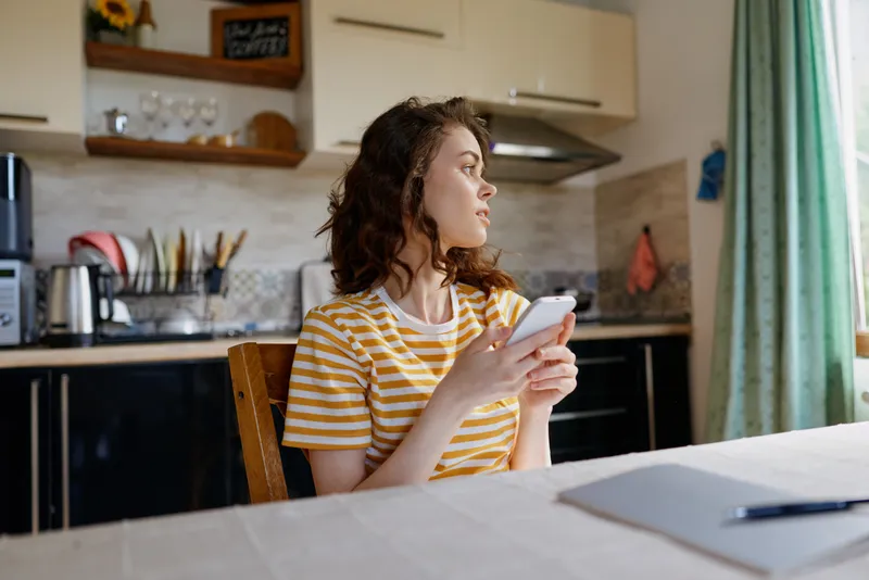 Young woman sits at a kitchen table, scrolling on her phone, striped yellow shirt in a cozy home environment, casual lifestyle and everyday moment, natural light and calm.