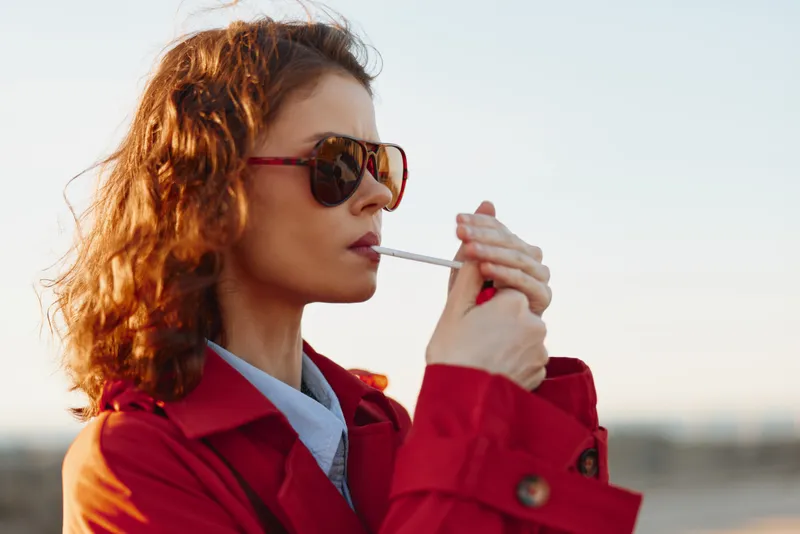 Young woman with curly hair wearing a red coat lights a cigarette outdoors with sunglasses on a clear day.
