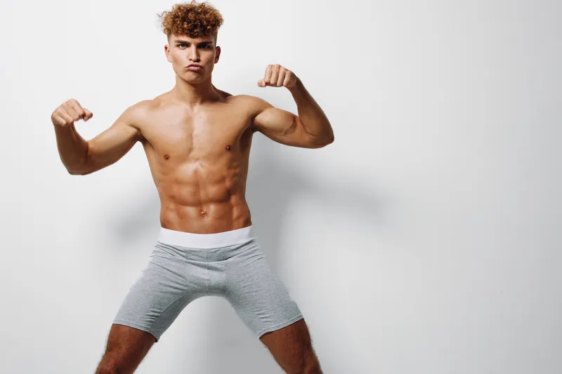 Young muscular man posing shirtless in gray shorts showing strong biceps and defined abs on white background for fitness, health and bodybuilding concept.