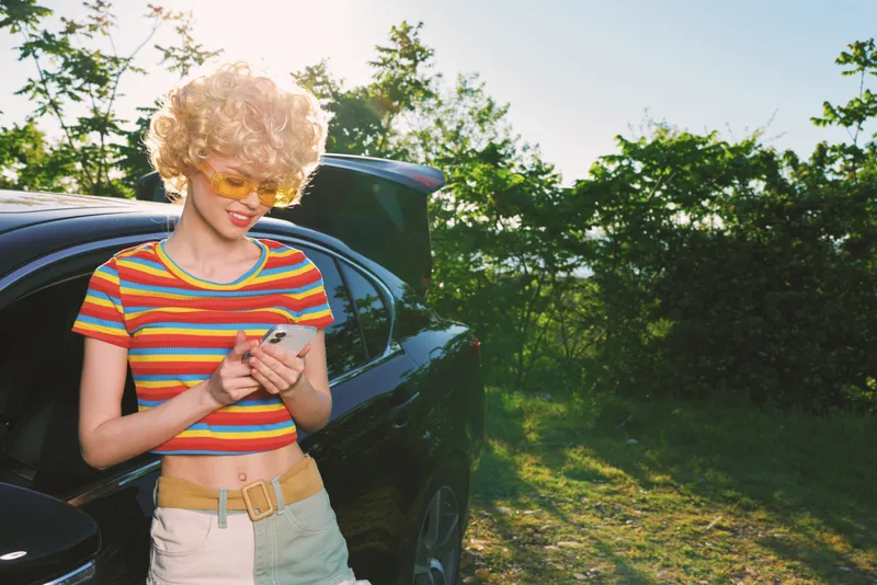 Smiling woman with curly hair wearing a striped crop top stands beside a car, looking at a phone. Outdoor lifestyle scene with retro color vibe and sunny warm lighting, candid and fresh.