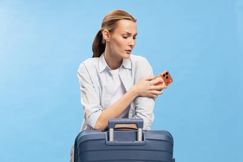 Young woman with suitcase looks at smartphone with focused expression. Travel concept with blue background, casual outfit, and preparation for trip. Modern lifestyle and communication theme.