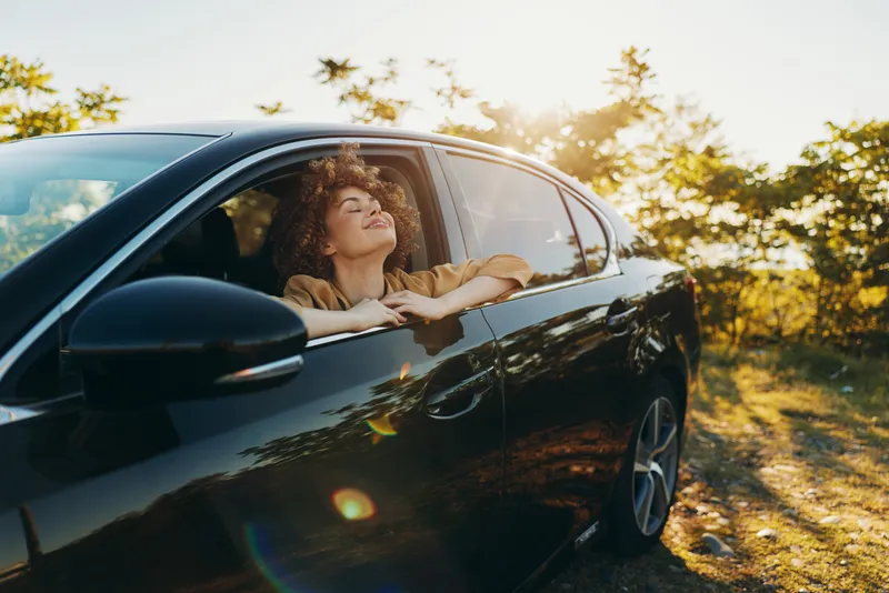 Smiling woman leaning from a car window in a sunlit countryside, enjoying a carefree moment. Outdoor lifestyle, retro film color, warm light creates a relaxed mood. for a nostalgic feel.