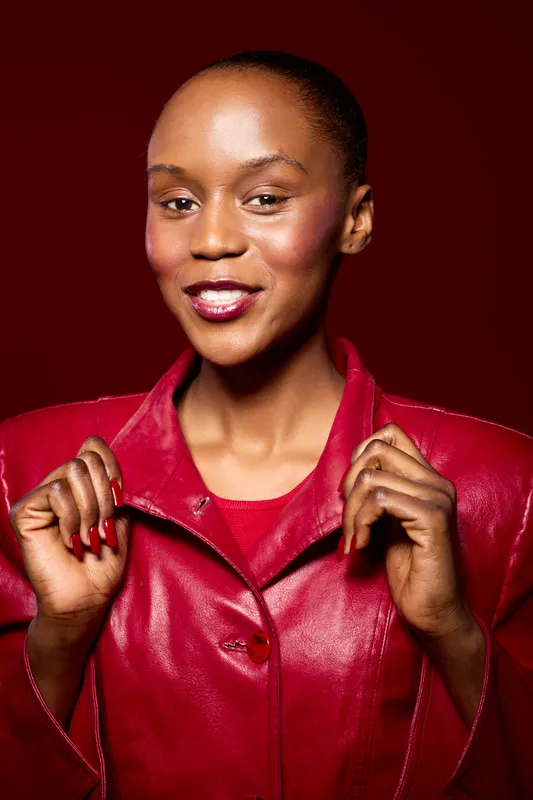 Confident black woman with dark skin wearing a red leather jacket smiles while holding the collar. Studio portrait on red background. Concept of fashion, beauty, and empowerment.