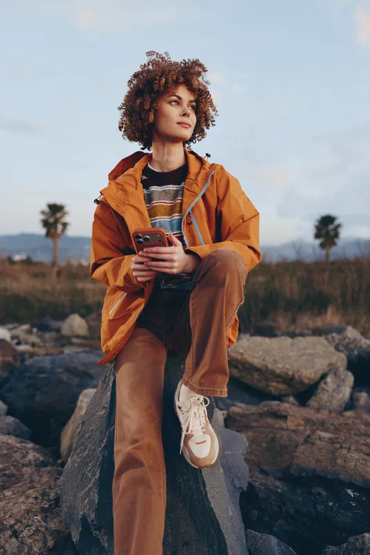 Woman lifestyle outdoors holding smartphone, wearing orange jacket, curly hair blowing slightly, relaxed pose on rocky landscape, golden hour sun, natural colors, calm mood, soft breeze