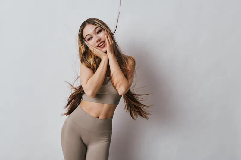 Young woman in beige sportswear posing with hands on face smiling. Fitness model with long hair in studio on light background for health and active lifestyle.