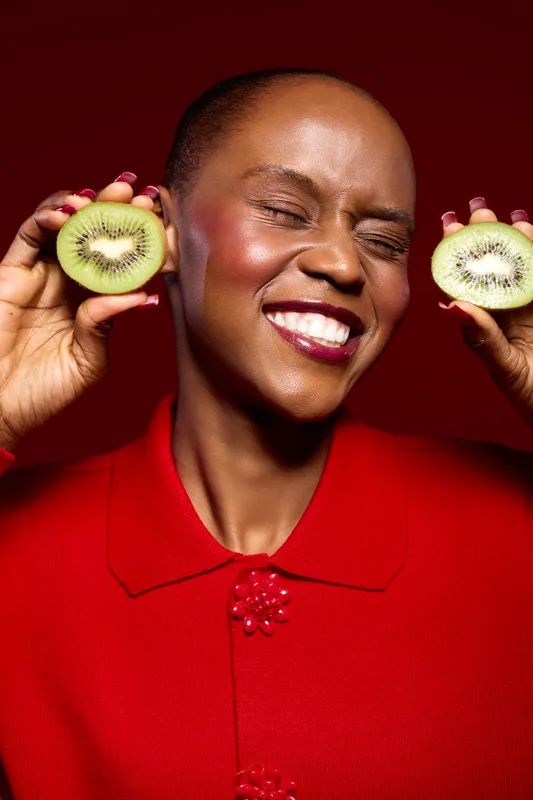 Smiling dark skinned African woman holds two kiwi halves near face with eyes closed. Bright red outfit contrasts with fresh green fruit. Concept for healthy lifestyle and natural beauty.