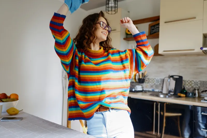 Joyful woman indoors dancing in a striped sweater, bright kitchen setting, natural light, casual home lifestyle moment, playful pose, vibrant colors, relaxed mood, with cheerful energy and everyday