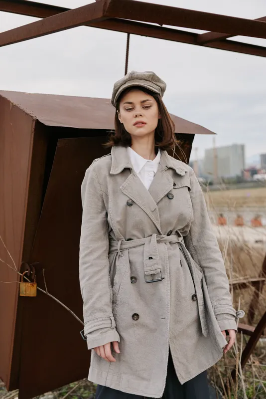 Young woman in textured coat and matching hat stands outdoors near metal structure with urban buildings in background. Fashion, autumn style, and casual mood concept.