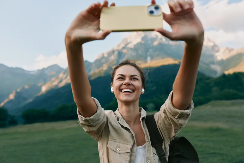 Young woman snaps a selfie outdoors on a sunlit mountain meadow, smiling as she frames the shot with a beige shirt and backpack.