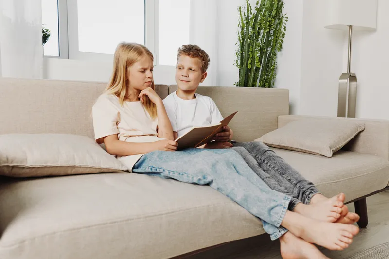 Two children sitting on a beige sofa reading a book together in a bright living room with natural light and plants in the background.