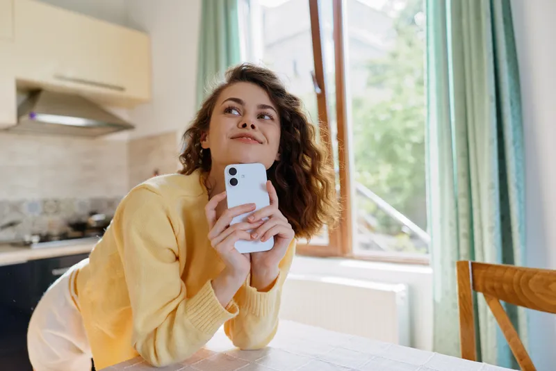 Young woman at home, casual lifestyle, sits at kitchen table with smartphone, warm sweater, natural light from window, cheerful relaxed mood, candid moment of everyday life.