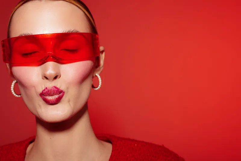 Close-up of a woman with red lips and red transparent glasses making a kiss face on a red background. Concept of fashion, beauty, and bold makeup with vibrant colors.