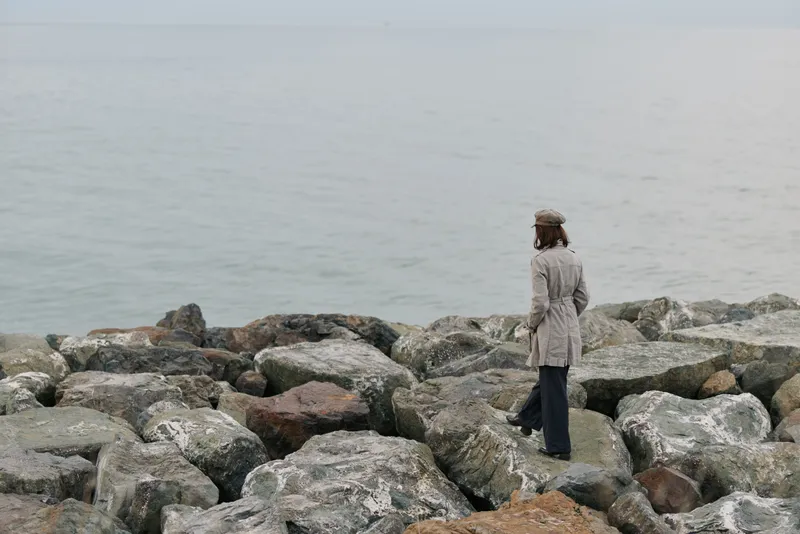 Woman in coat and hat stands on rocky shore looking at calm sea. Overcast weather creates peaceful and contemplative mood. Concept of solitude, nature, and reflection.