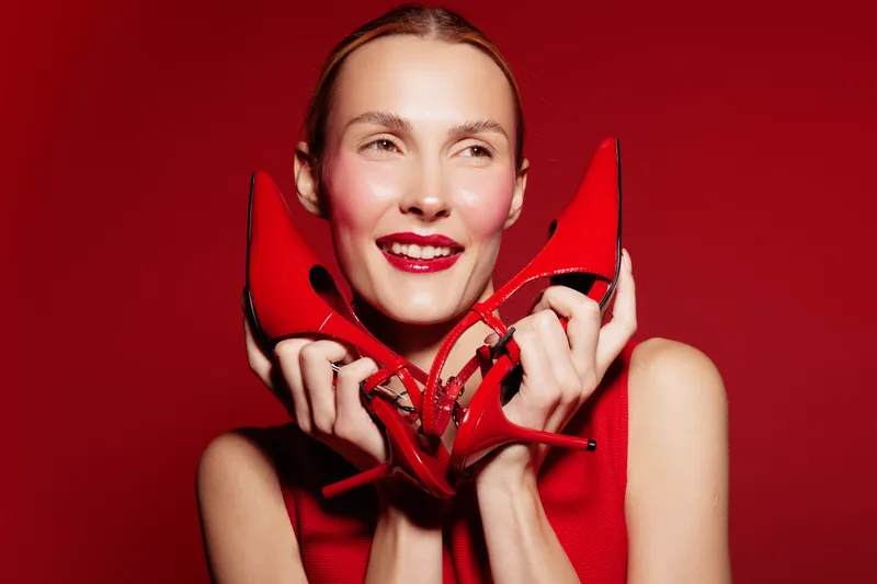 Smiling woman in red dress holds red high heels near face against red background. Concept of fashion, style, beauty, and confident female model with vibrant makeup and elegant accessories.