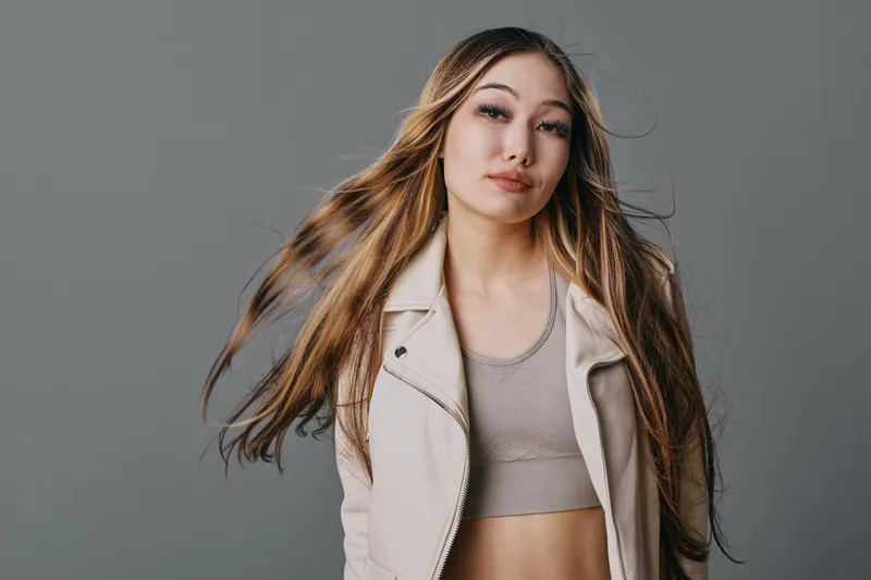 Young woman with long hair wearing beige jacket and crop top posing against gray background with natural expression and flowing hair in studio portrait.