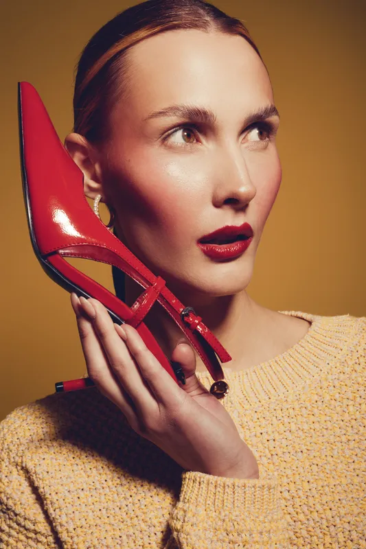 Stylish woman with red lipstick holds red high heel shoe near face, wearing yellow sweater. Fashion concept with bold makeup and elegant accessory on warm brown background.
