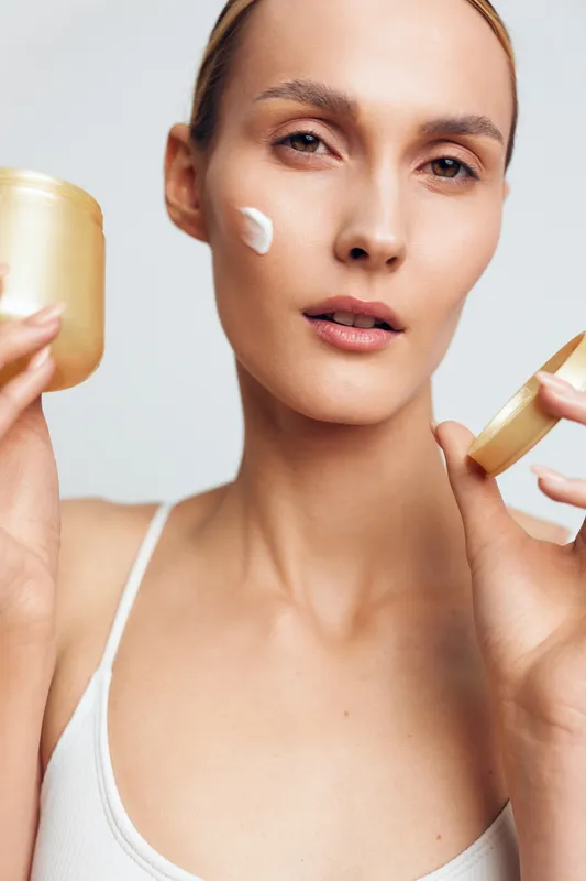 Young woman applies moisturizing cream on face holds open jar skincare product shows healthy skin beauty routine isolated on light background natural look and fresh appearance.